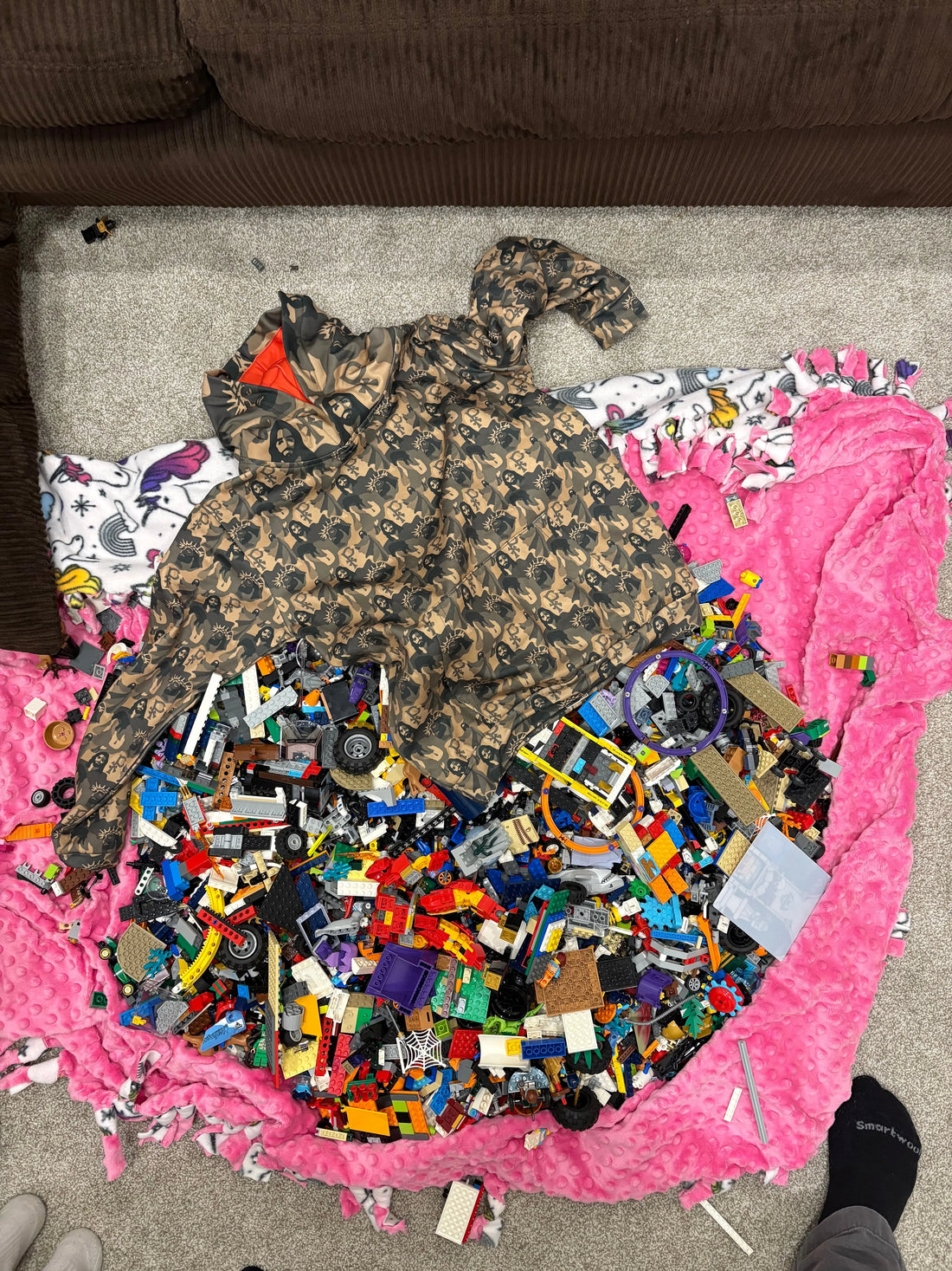 An image of an unfolded Jesus camo hoodie on the floor amidst colorful legos and a pink blanket. 